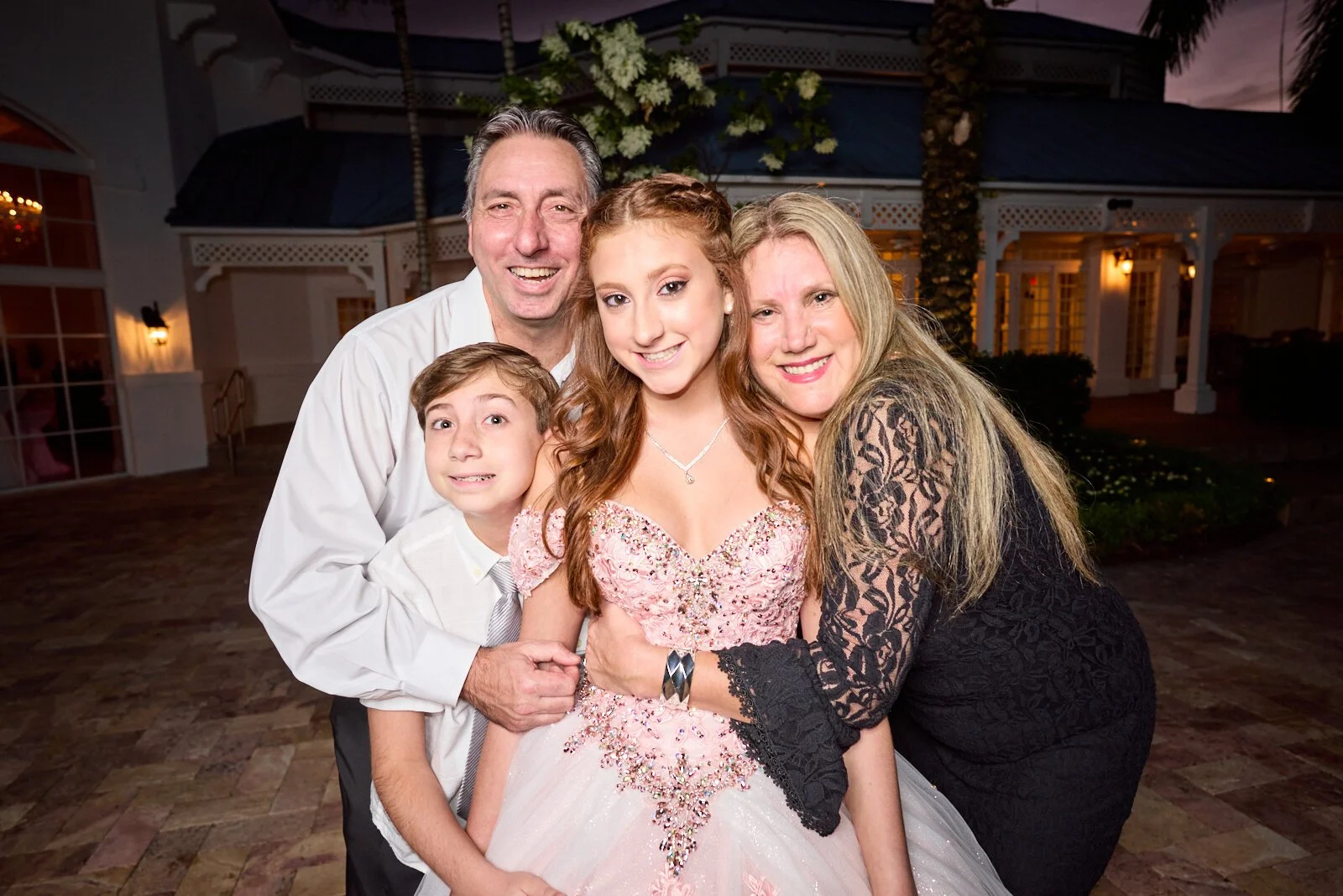 Family of four posing together outdoors during evening at a celebration, with a house and yard in the background.