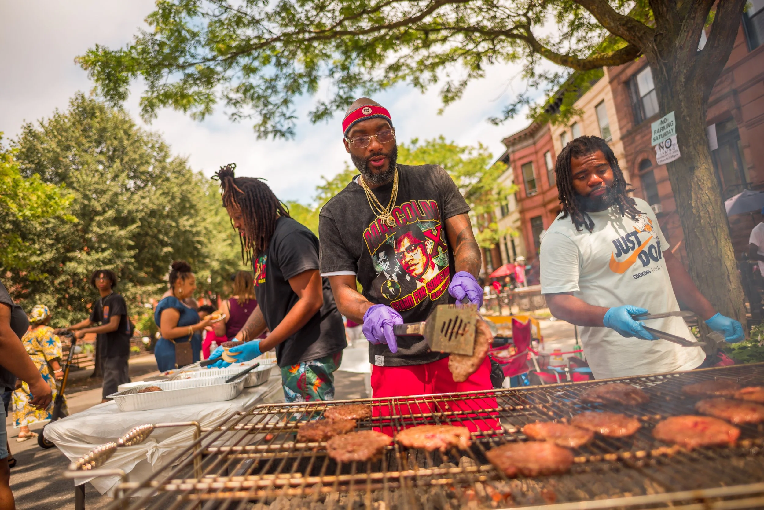 Three men in colorful casual clothes grilling and serving barbecue at an outdoor community event in a park with many people and trees.