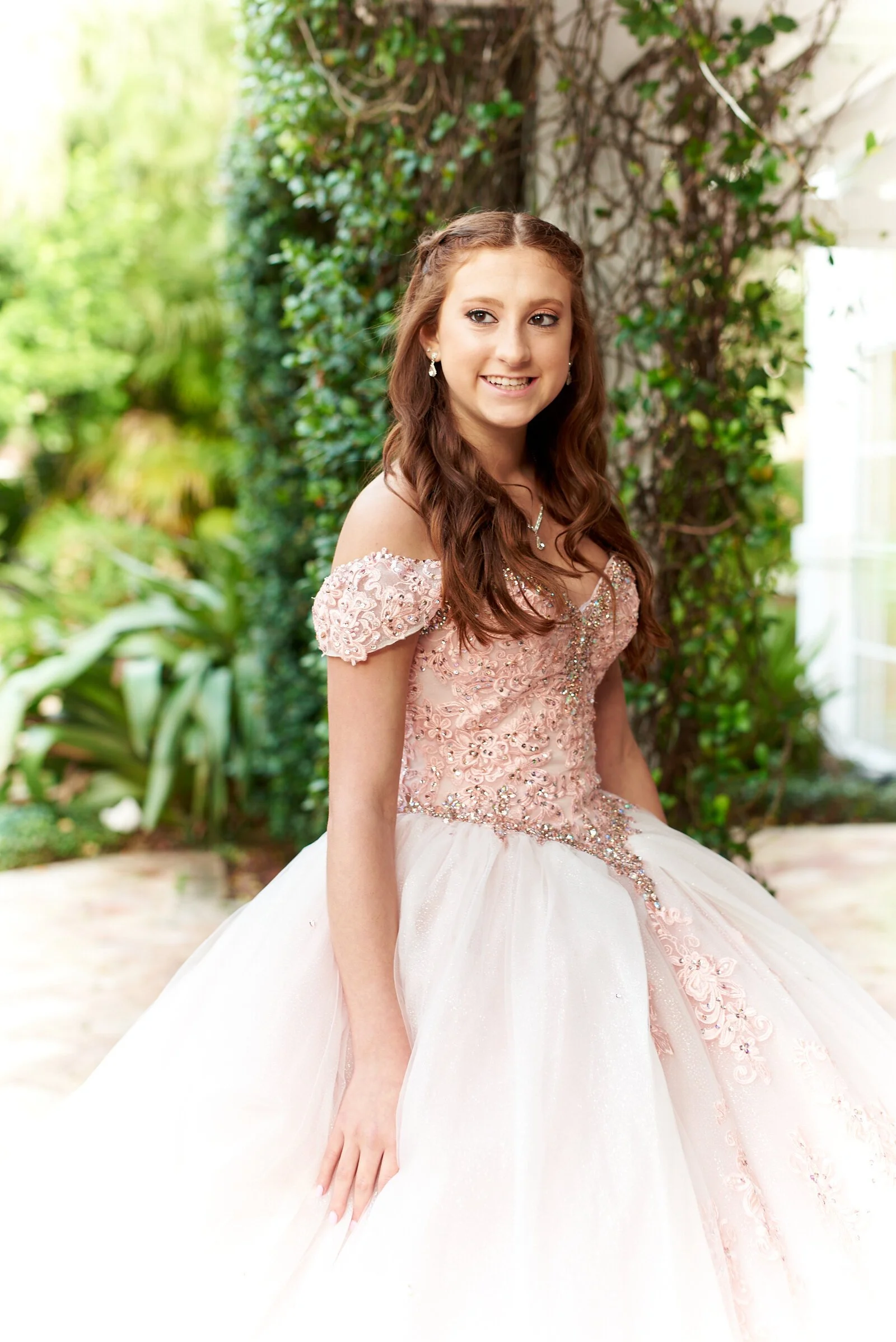 A young woman in an ornate pink and white ball gown with lace details, standing outdoors in front of greenery, smiling.