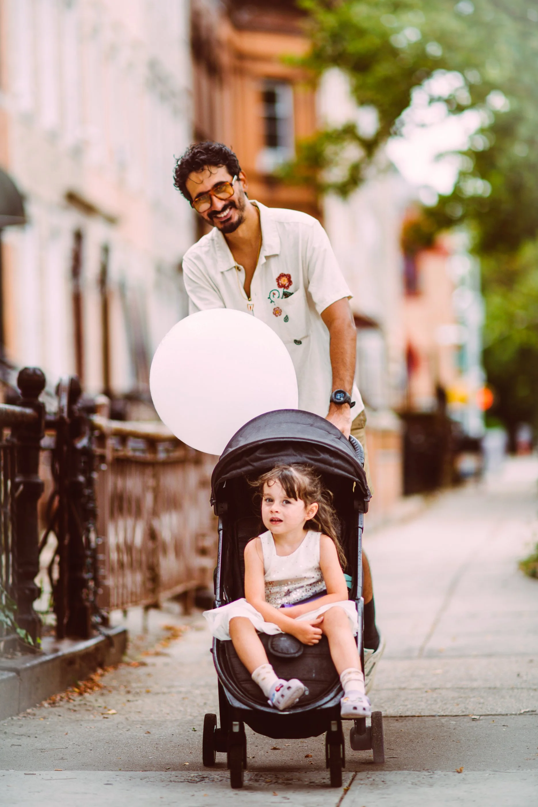 A man with glasses and a beard pushes a stroller with a young girl sitting inside, holding a white balloon, on a city sidewalk lined with trees and buildings.