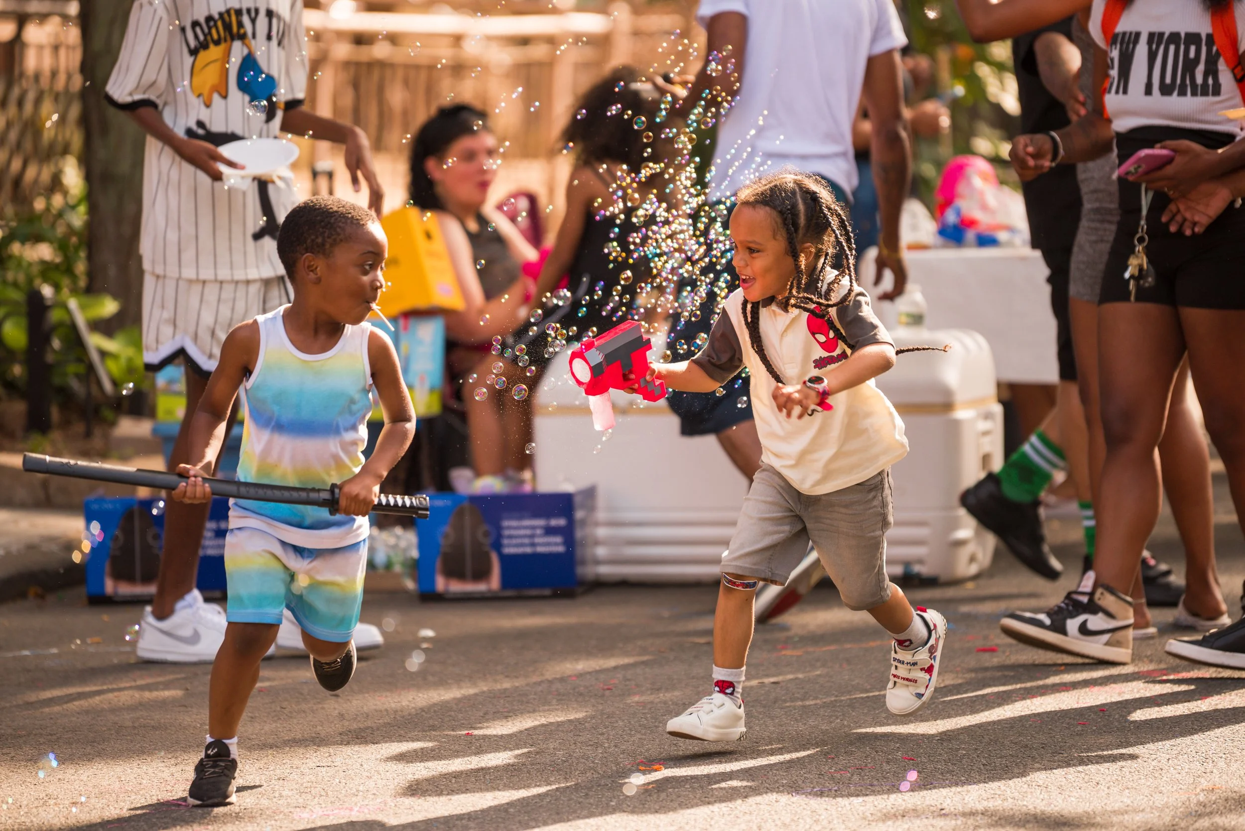 Children playing with bubbles and toy guns at a celebration or outdoor party, with adults and various items in the background.