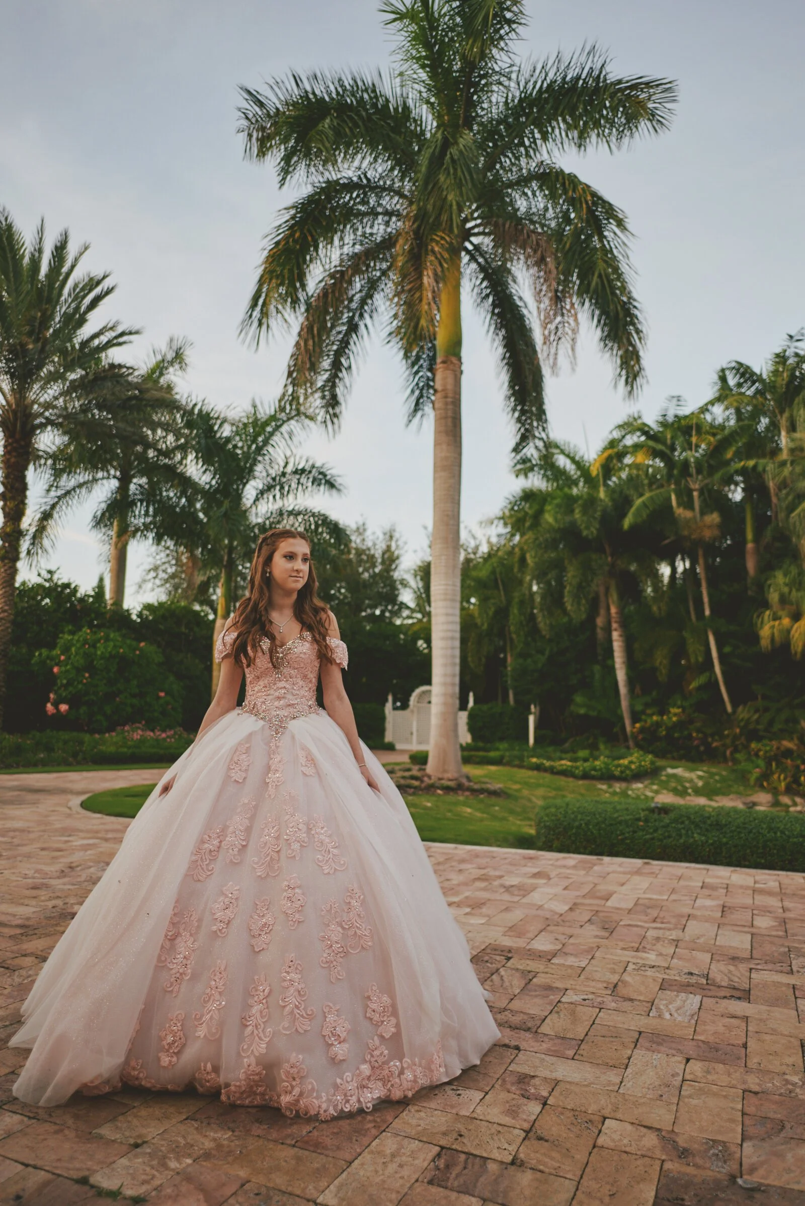 A young woman in a pink and white floral ball gown standing outdoors on a brick pathway, with palm trees and lush greenery in the background during daylight.