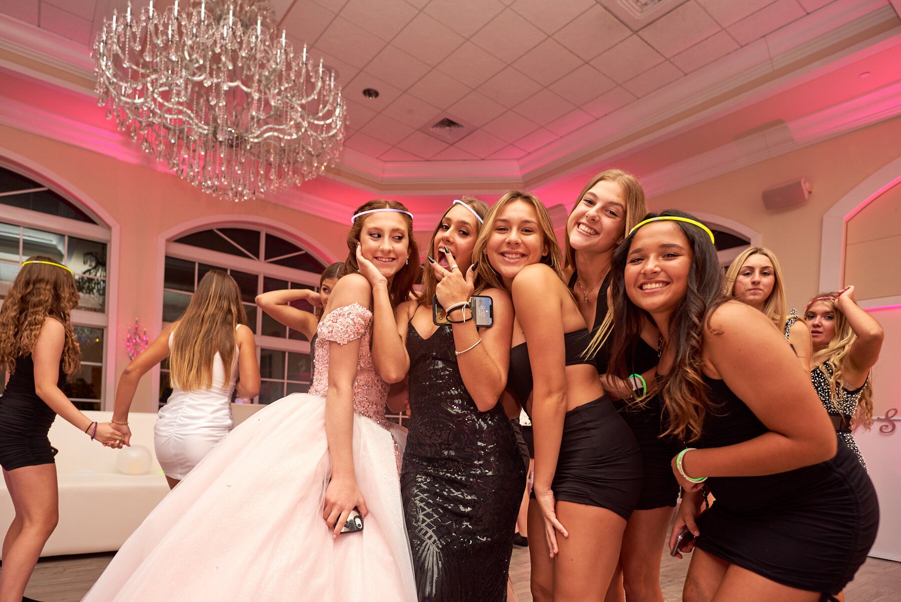 Group of young women at a party, dressed in elegant and casual clothes, smiling and posing for a photo under a large chandelier in a decorated venue with pink lighting.