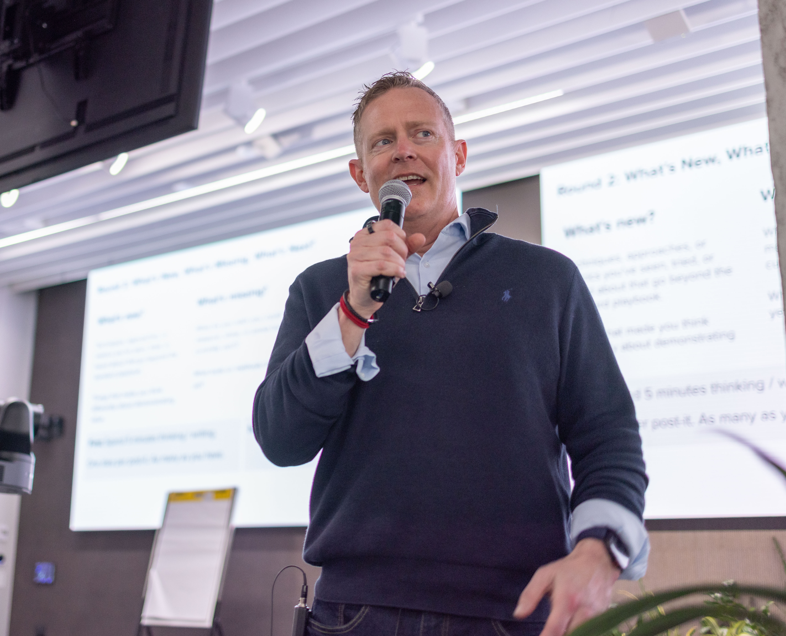A man with short hair, wearing a dark sweater over a collared shirt, is holding a microphone and speaking. There is a large screen with text behind him, and the setting appears to be a conference or presentation room.