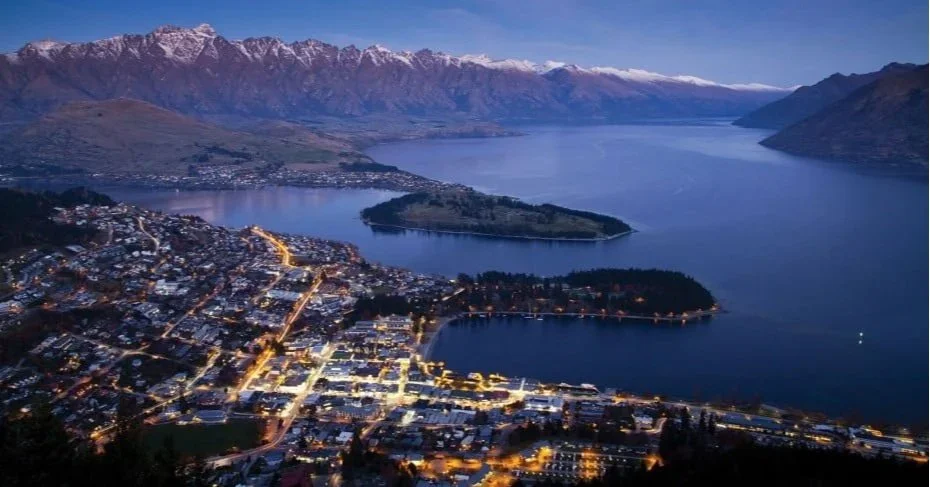 Aerial view of a city by a lake with snow-capped mountains in the background during twilight.
