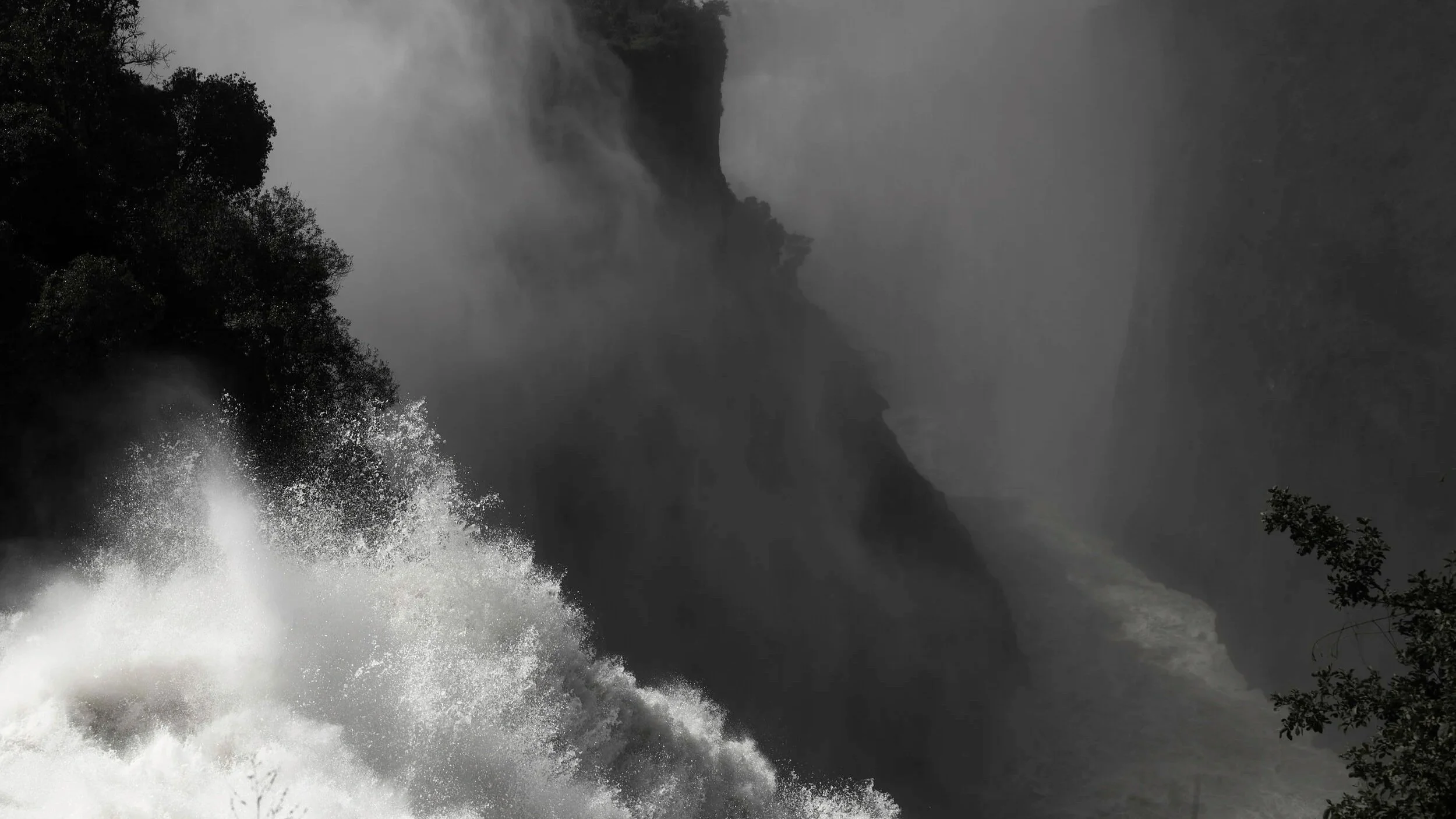A powerful waterfall crashing down on rocks with mist and spray, surrounded by trees in a misty, cloudy atmosphere.