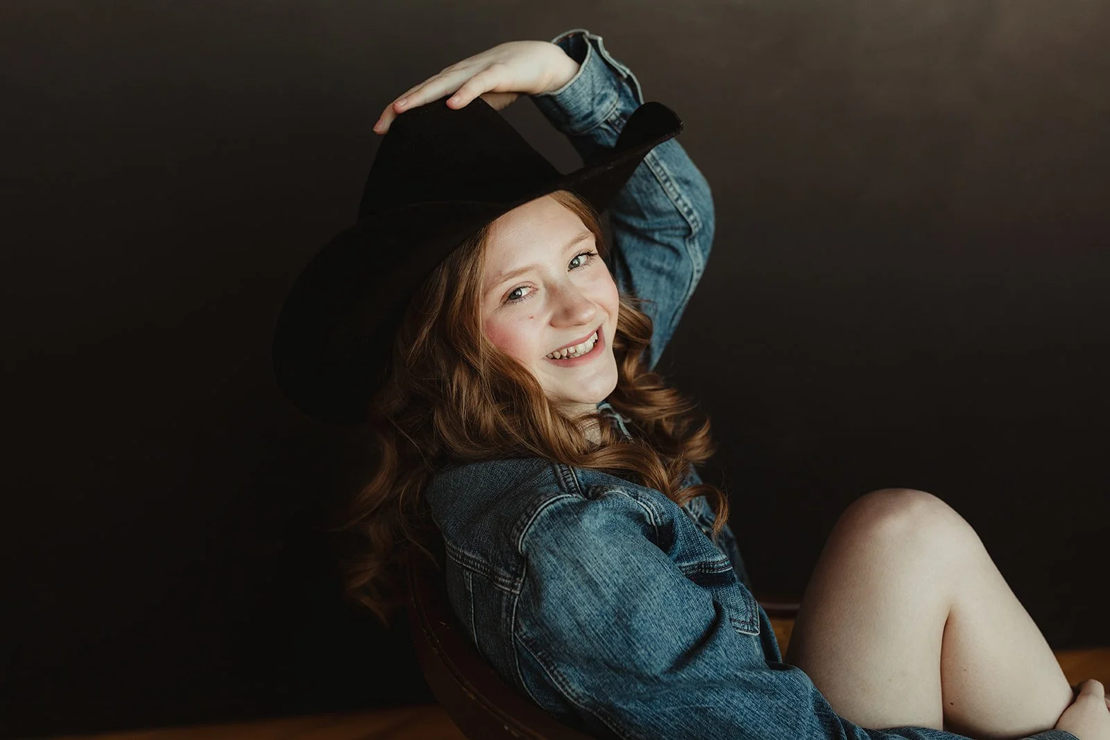A young woman with red hair smiling, wearing a black wide-brimmed hat and denim jacket, sitting against a black background.