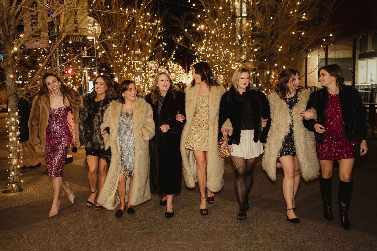 A group of women wearing party dresses and coats walking arm-in-arm down a city street decorated with string lights at night.