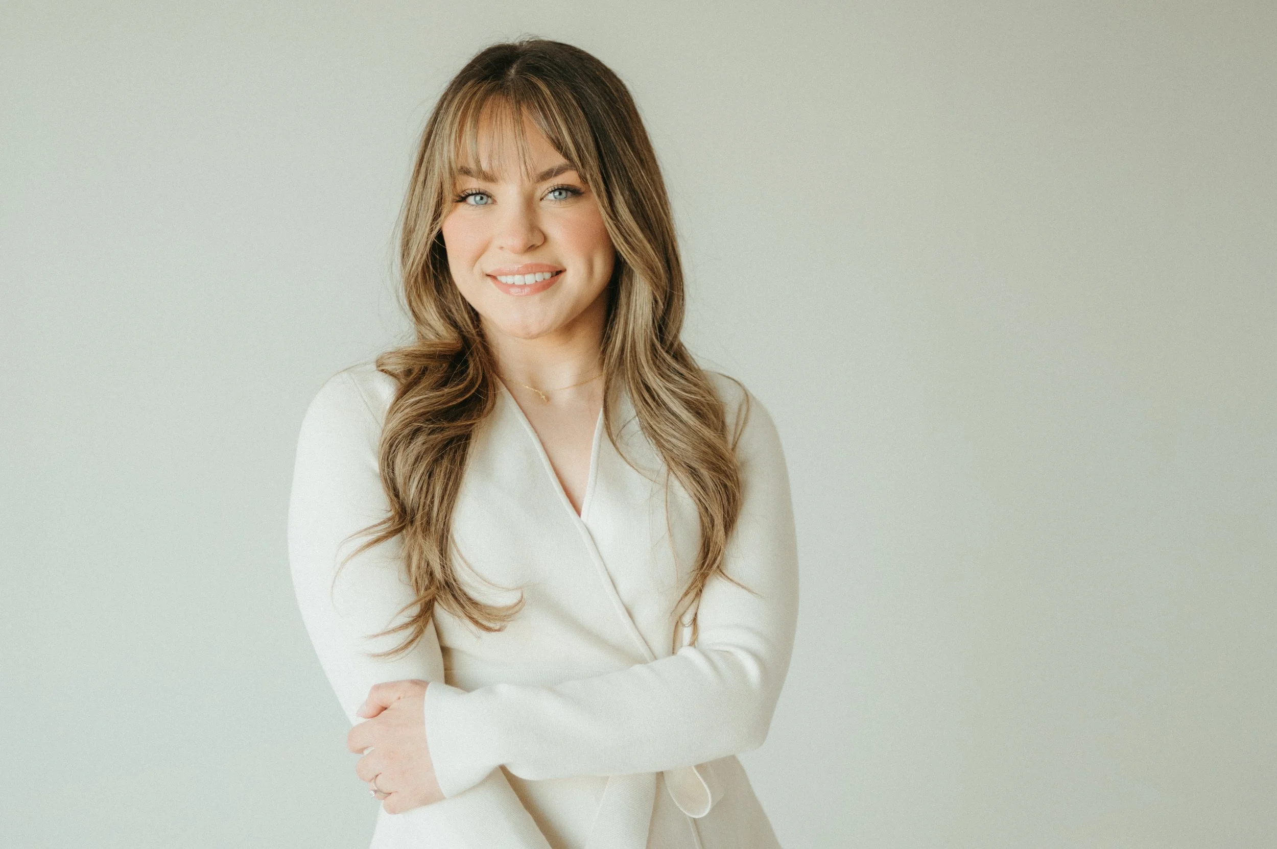 A young woman with long wavy brown hair and blue eyes, smiling and wearing a white blazer, standing against a plain light background.