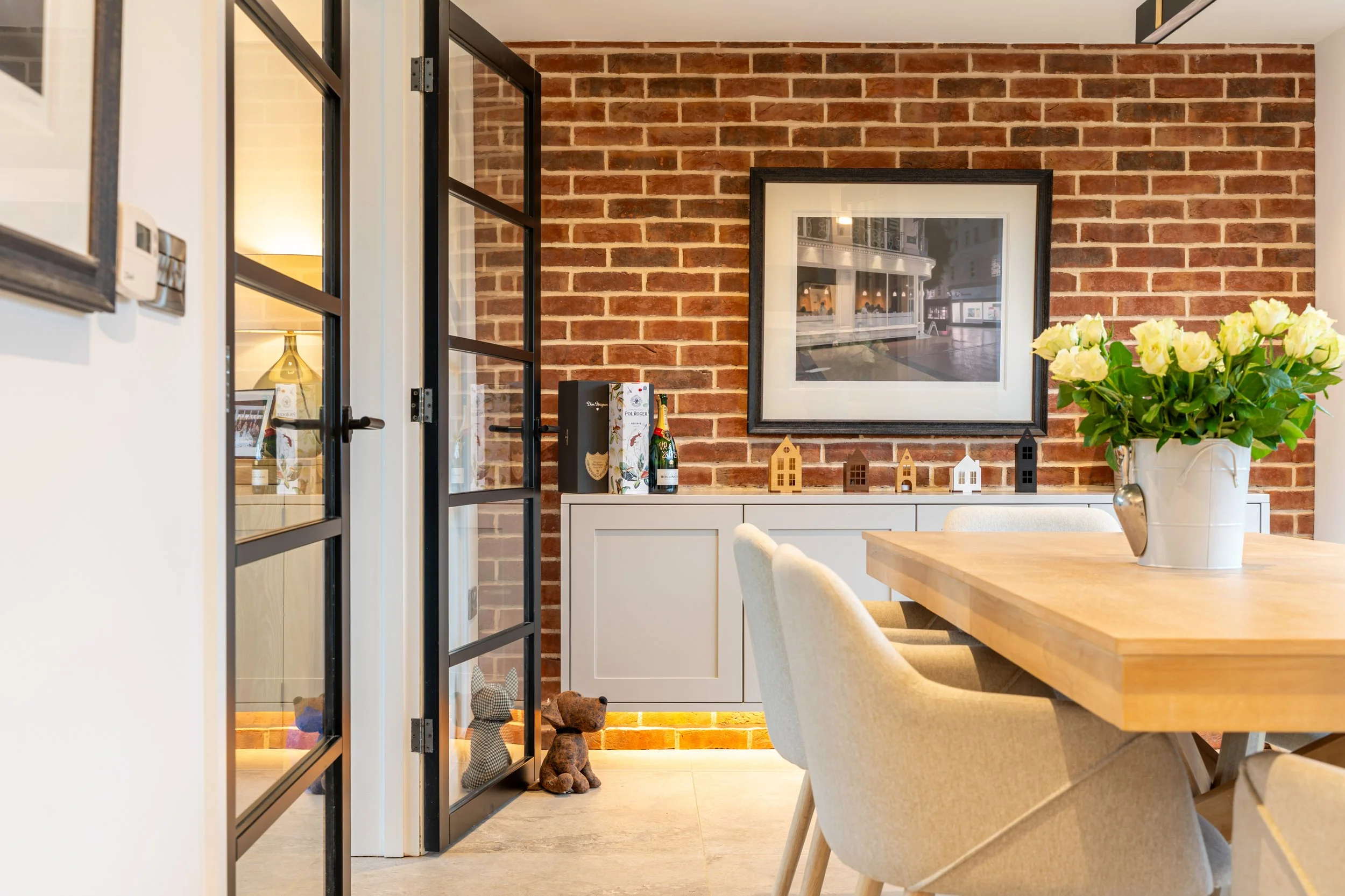 A glimpse into a dining or living area featuring a brick wall, a large framed photograph of a city street scene, a white cabinet with decorative items including a bottle, a small house-shaped ornament, and plush teddy bears. There is a wooden dining table with light-colored upholstered chairs, and a potted plant with white flowers on the table.