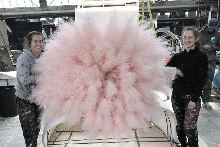 Two women standing on either side of a large pink fluffy feather sculpture in an indoor industrial space.