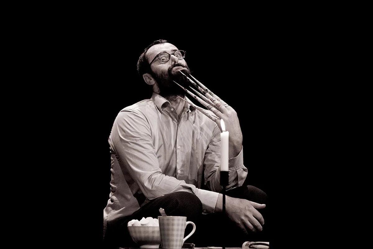 Ein Mann mit langen, spitzen Fingernägeln sitzt im Schneidersitz. Er trägt ein hellblaues Hemd und hält eine Kerze in der Hand. Auf dem Tisch vor ihm stehen eine Tasse und eine Schüssel mit Zuckerstückchen.