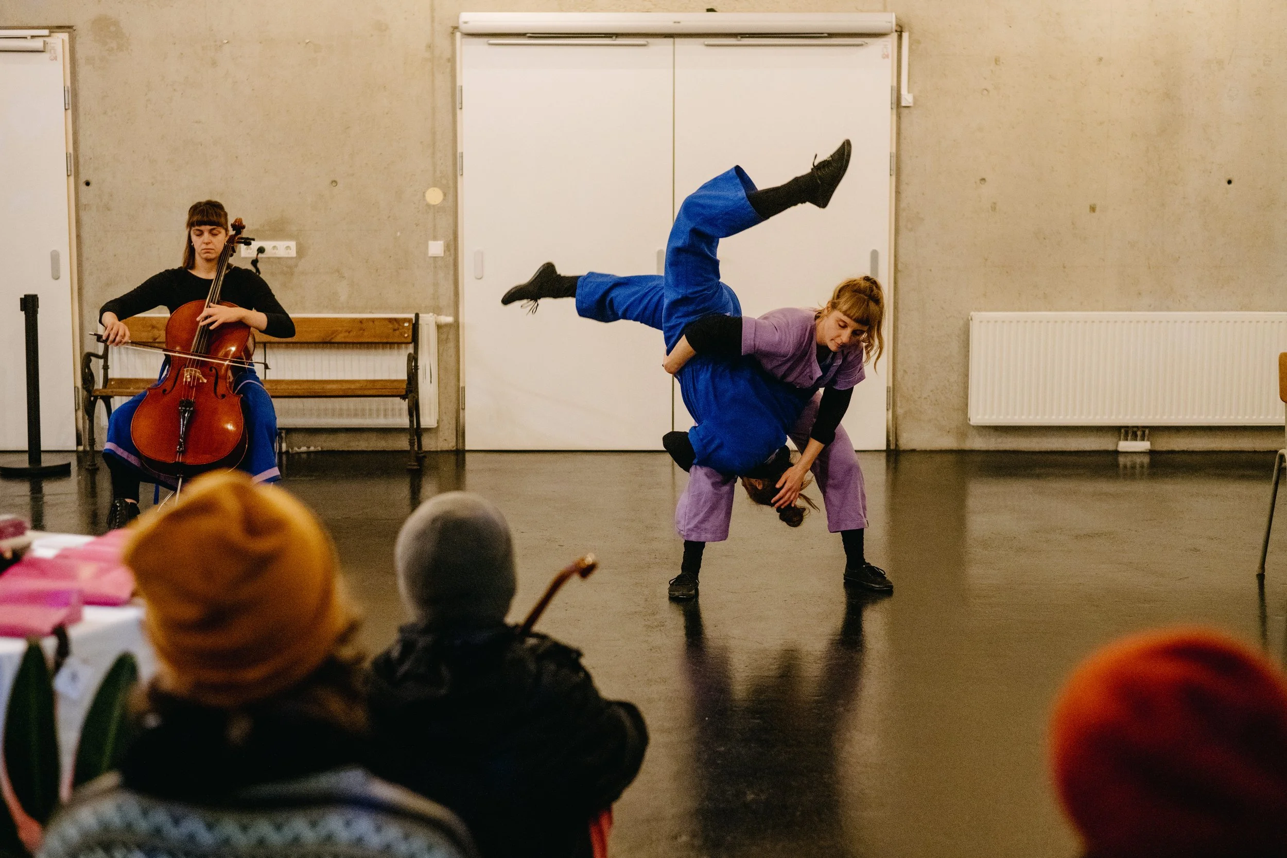 Zwei Tänzer*innen in lila und blau führen einen Partner*innenakrobatik-Trick auf. Im Hintergrund sitzt eine Cellistin. Zuschauer*innen sind teilweise sichtbar.