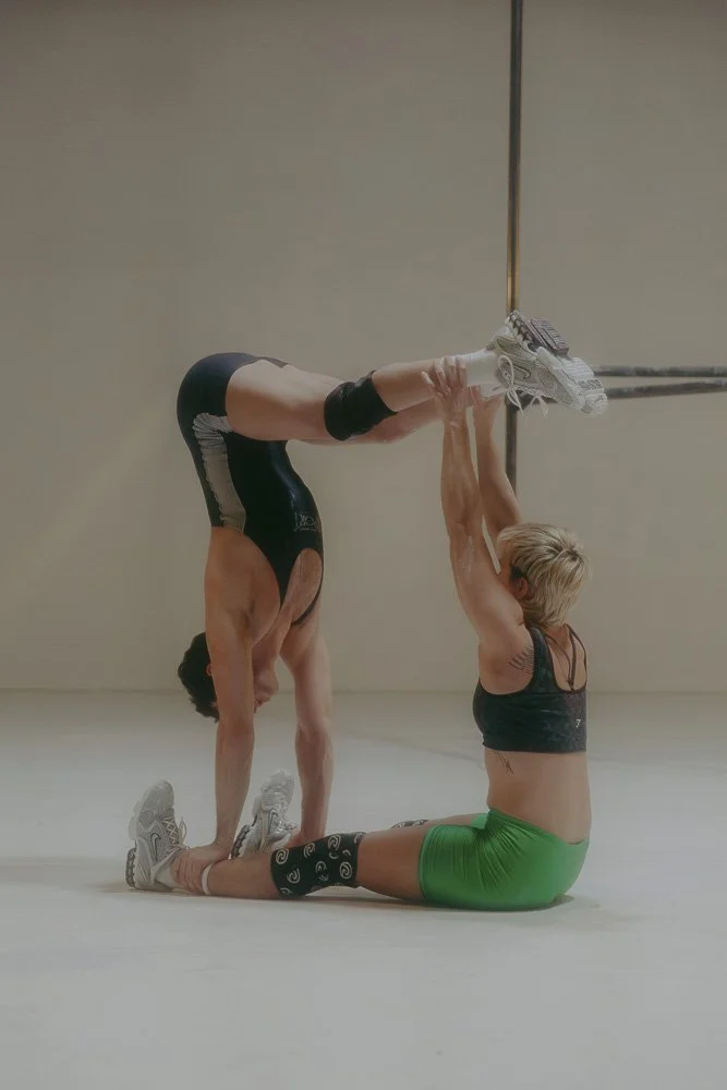 Zwei Personen in Sportkleidung führen eine akrobatische Übung aus. Eine Person steht auf den Händen, während die andere sie an den Beinen hält. Die erste Person trägt einen schwarzen Anzug, die zweite Person ein grünes Sportoutfit.