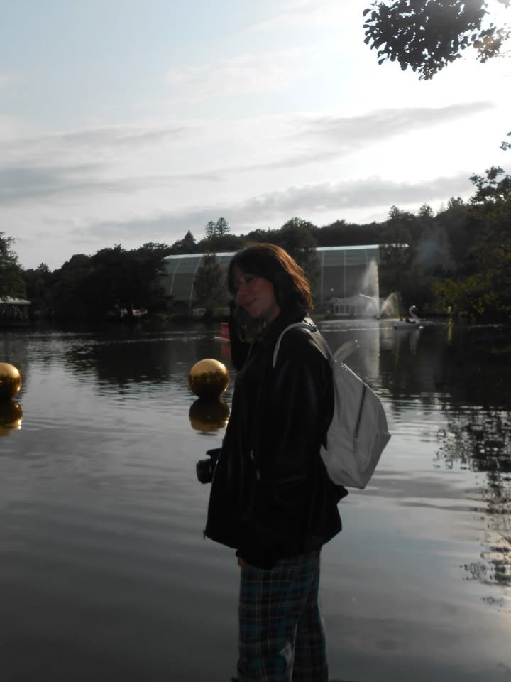 A woman standing by a river with trees and a fountain in the background, wearing a black jacket, plaid pants, and a white backpack, during sunset.