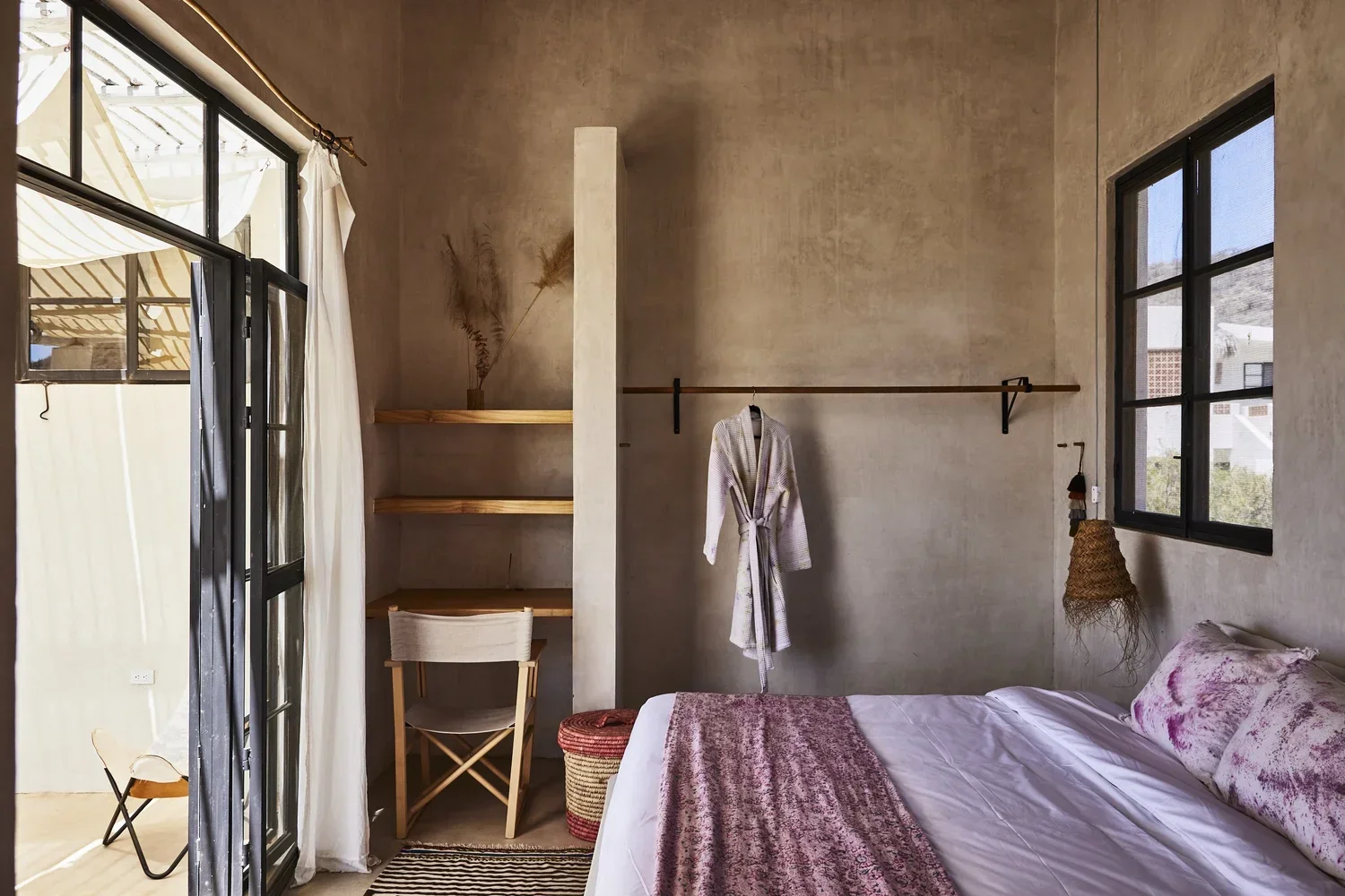 A cozy bedroom with a large bed covered in white sheets and pink patterned pillows and blanket, beside a window with black frames, a vanity desk with a chair, a wall-mounted wooden rod with a bathrobe hanging, and a shelving unit with dried plants, natural light coming through the windows.