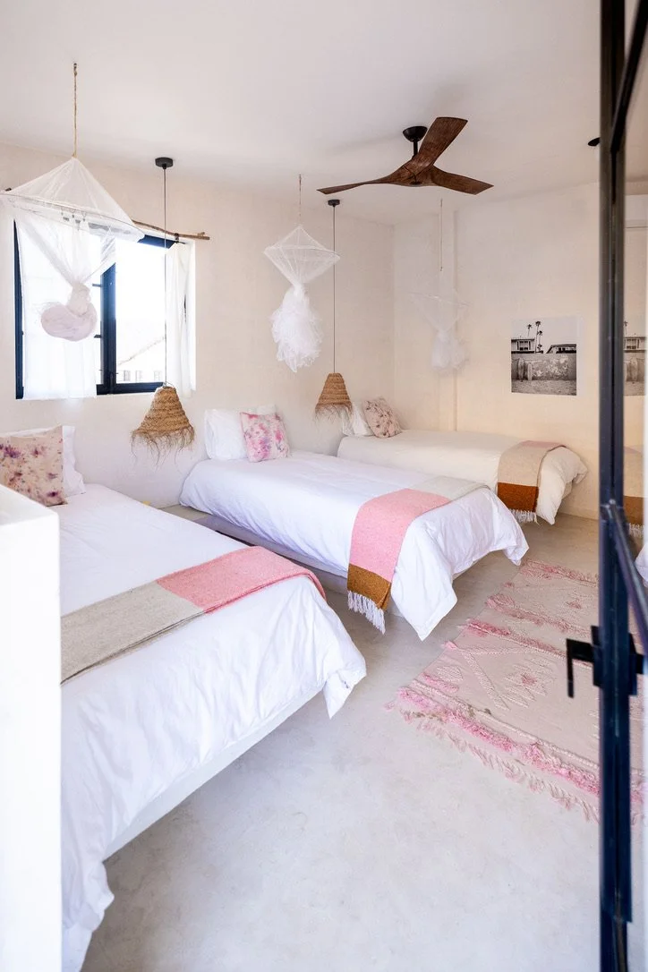 A bedroom with three single beds, white bedding, pink and beige throws, floral pillows, hanging woven lampshades, a ceiling fan, a window with white curtains, and a pink and beige rug.
