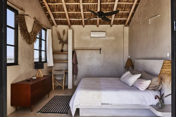A cozy bedroom with a minimalist design. It features a large bed with white bedding, two bedside lamps with woven shades, a simple wooden dresser, and a small desk with a chair. The walls are unfinished concrete, and there are large windows with white curtains letting in natural light. The ceiling is made of exposed wooden beams with a ceiling fan. Decor includes dried plants and minimal accessories.