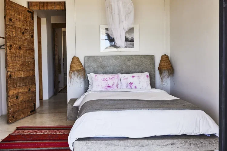 Cozy bedroom with a gray upholstered bed, pink and white pillows, two woven wall lamps, a black and white framed photo, a white canopy above the bed, a wooden sliding door, and a red striped rug.
