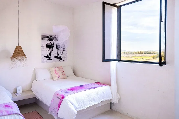 A bright bedroom with an open window showing a scenic view of fields and sky, white walls, a double bed with white bedding and pink accents, a painting of zebras, and a hanging woven light fixture.