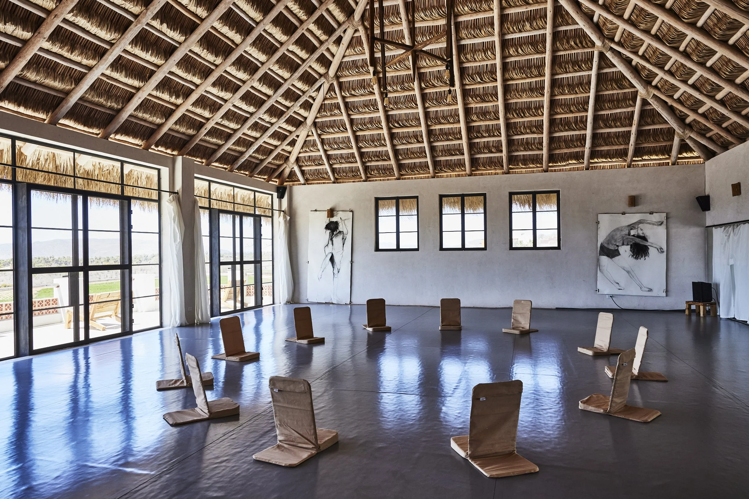 A spacious room with a high thatched roof and large windows. Wooden chairs are arranged on the floor in a circle. The walls feature modern black and white artwork of dancers, and the room has natural light streaming in.