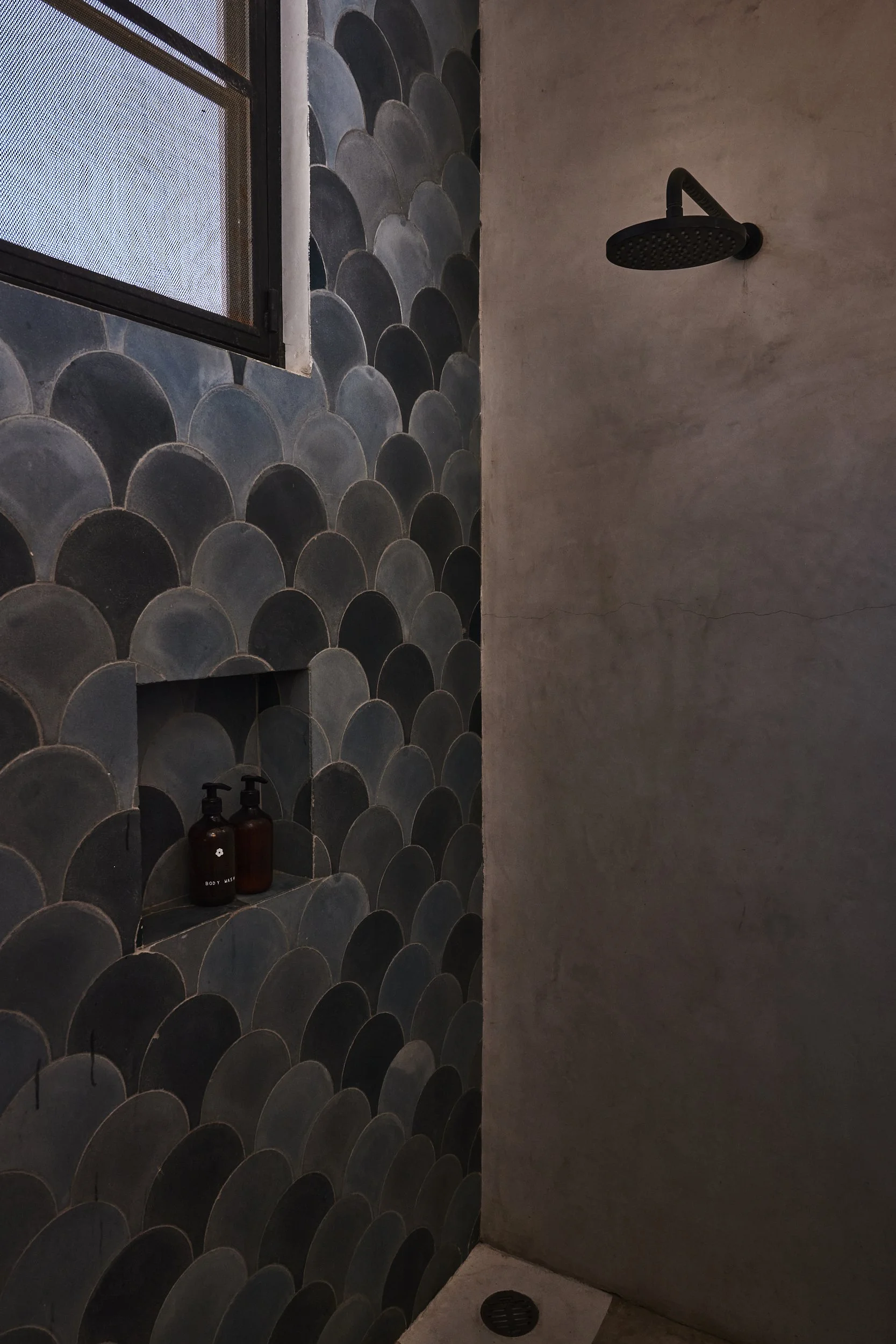 A shower area with a black round showerhead mounted on a beige wall. The wall on the left side is decorated with dark, scalloped, tile-like patterns. There is a small recessed shelf in the patterned wall holding two amber bottles labeled 'body wash' and 'shampoo'. A small window with a screen is visible at the top of the patterned wall.