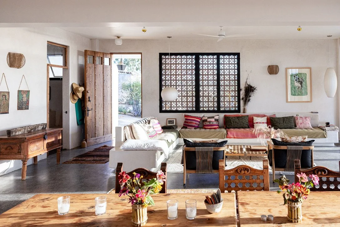 Living room and dining area with a wooden table, floral arrangements, a large L-shaped couch, and various chairs, with a front door open to a garden.