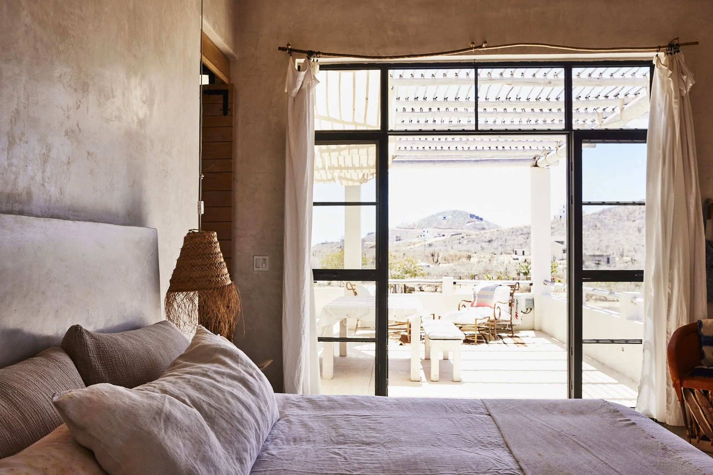 Bedroom with bed, beige pillows, wicker lampshade, sliding glass door leading to a terrace with outdoor seating, hills in the background.