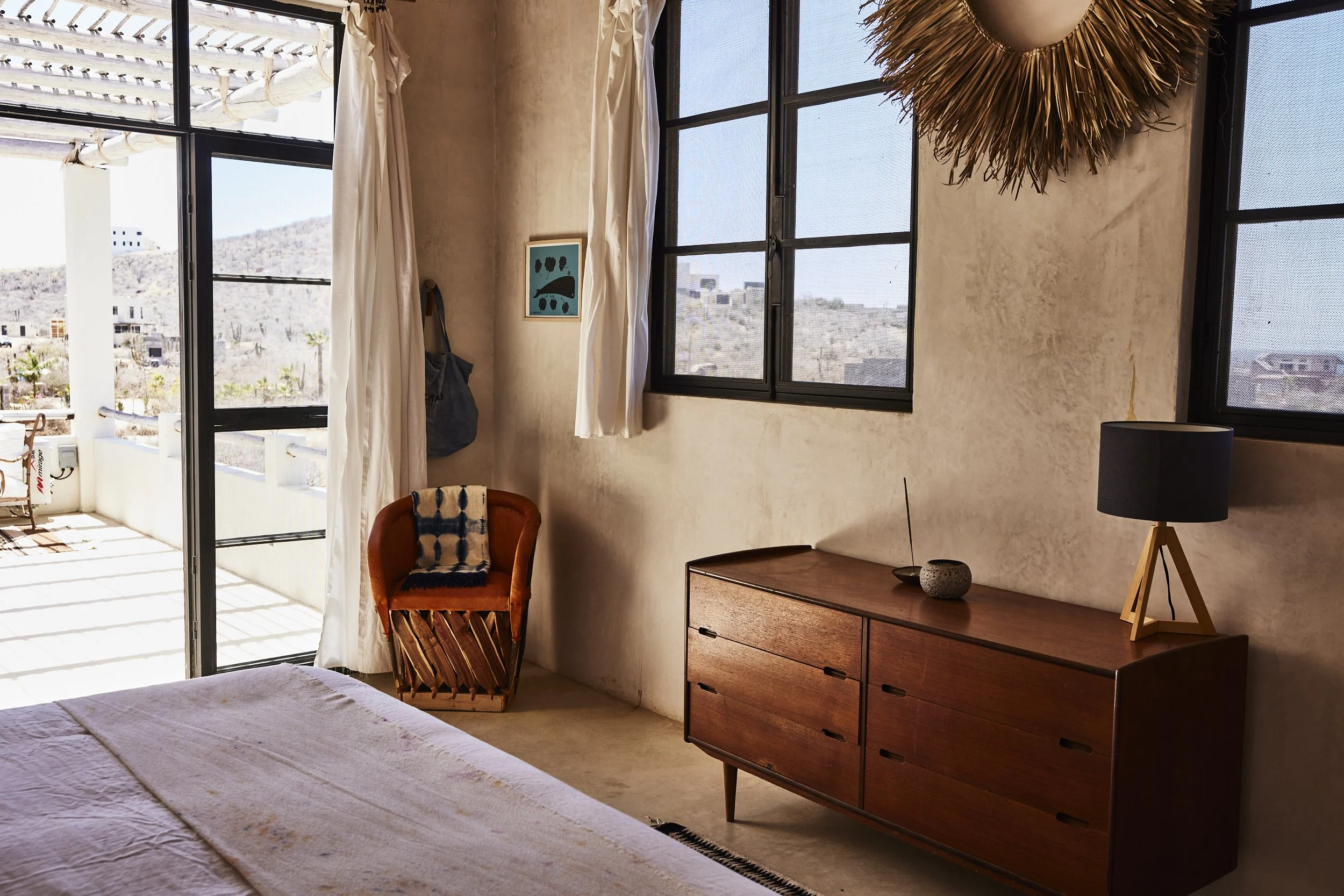A bedroom with a bed, wooden dresser with a lamp and decorative items, a chair with a pillow, and windows showing an outdoor mountain landscape, bright sunny day, light-colored walls, and minimalist decor.