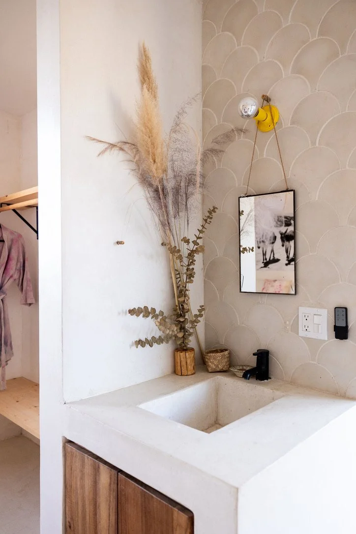 A cozy bathroom corner with a white concrete sink and wooden cabinet, decorated with tall dried pampas grass and eucalyptus in a wooden vase, on a built-in ledge. A yellow wall sconce with an exposed bulb illuminates a small mirror hanging on a patterned wall. There are electrical outlets and a remote on the wall, and a closet with a pink shirt and wooden hanger is partially visible on the left.