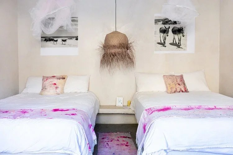 Two neatly made beds with white sheets and pink stains, decorated with pillows. Between them, there is a nightstand with a framed picture and a yellow candle. The wall above features black and white photographs of animals, a central hanging woven wall decoration with feathers, and white netting or fabric adornments.