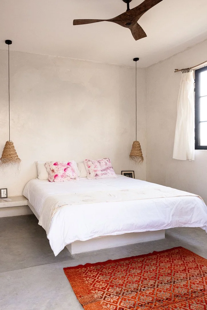 A minimalist bedroom with a white bed, pink and white pillows, hanging wicker lights, a white window with curtains, a small framed picture, and a red patterned rug.