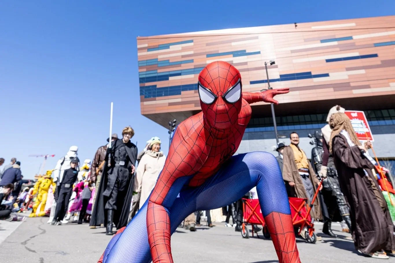 A photo of a person dressed up as Spiderman, posing in front of the copper facade of the BMO Centre on Stampede Park in Calgary's Culture and Entertainment District. There are other cosplayers in the background.