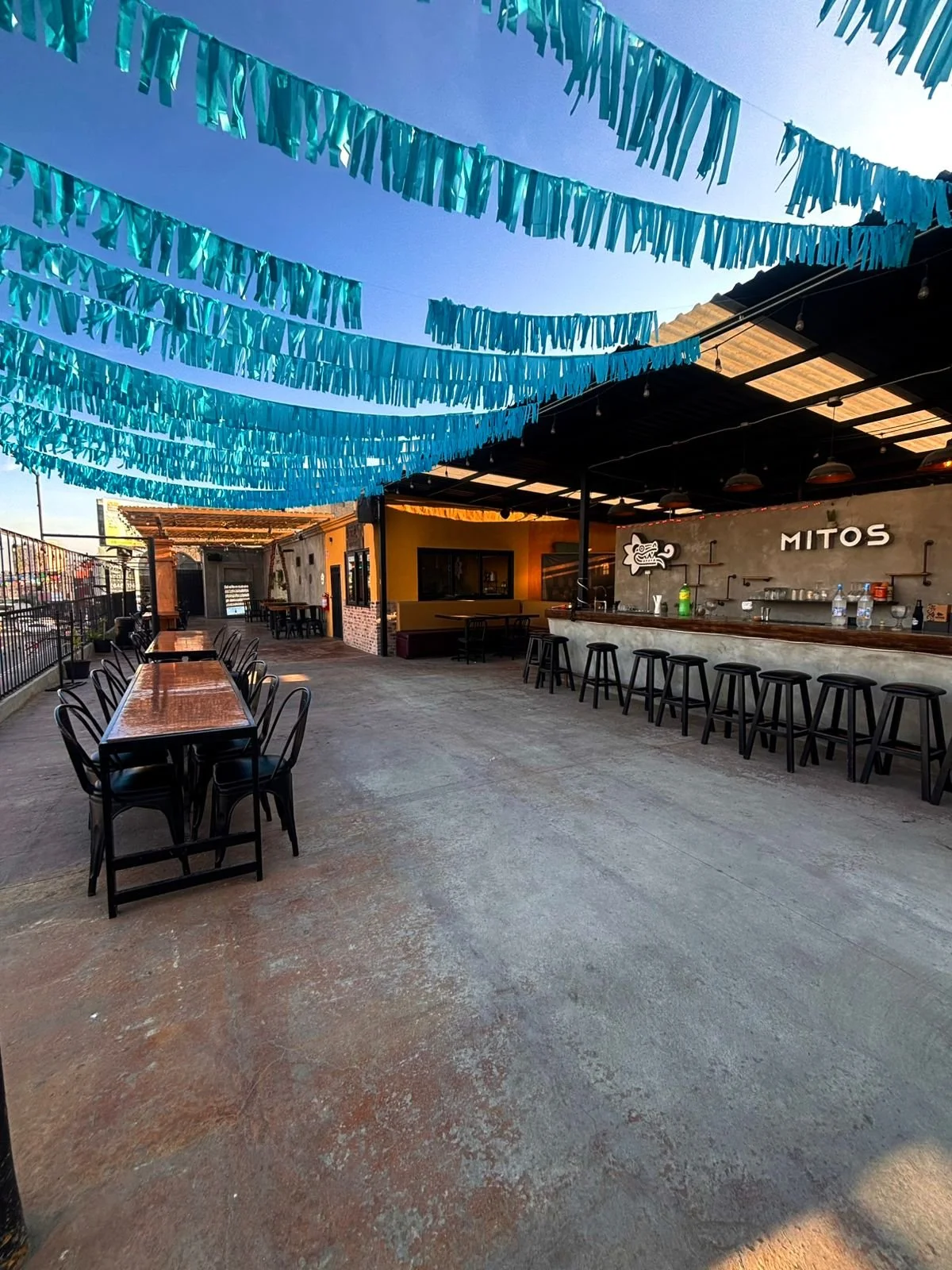 Outdoor bar area decorated with blue paper streamers, with high tables and bar stools, under a roof with string lights, and a sign that reads 'MITOS' on the back wall.