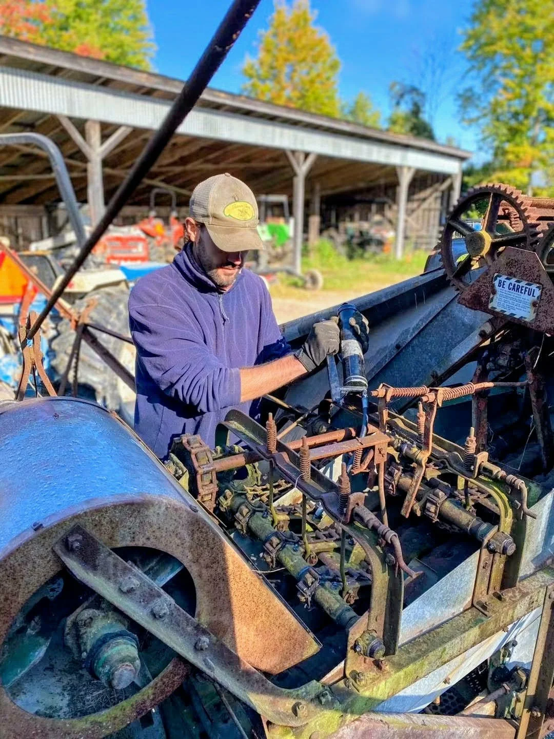 Man in a blue zip up fleece with a tan ball cap on working on a large piece of farm machinery