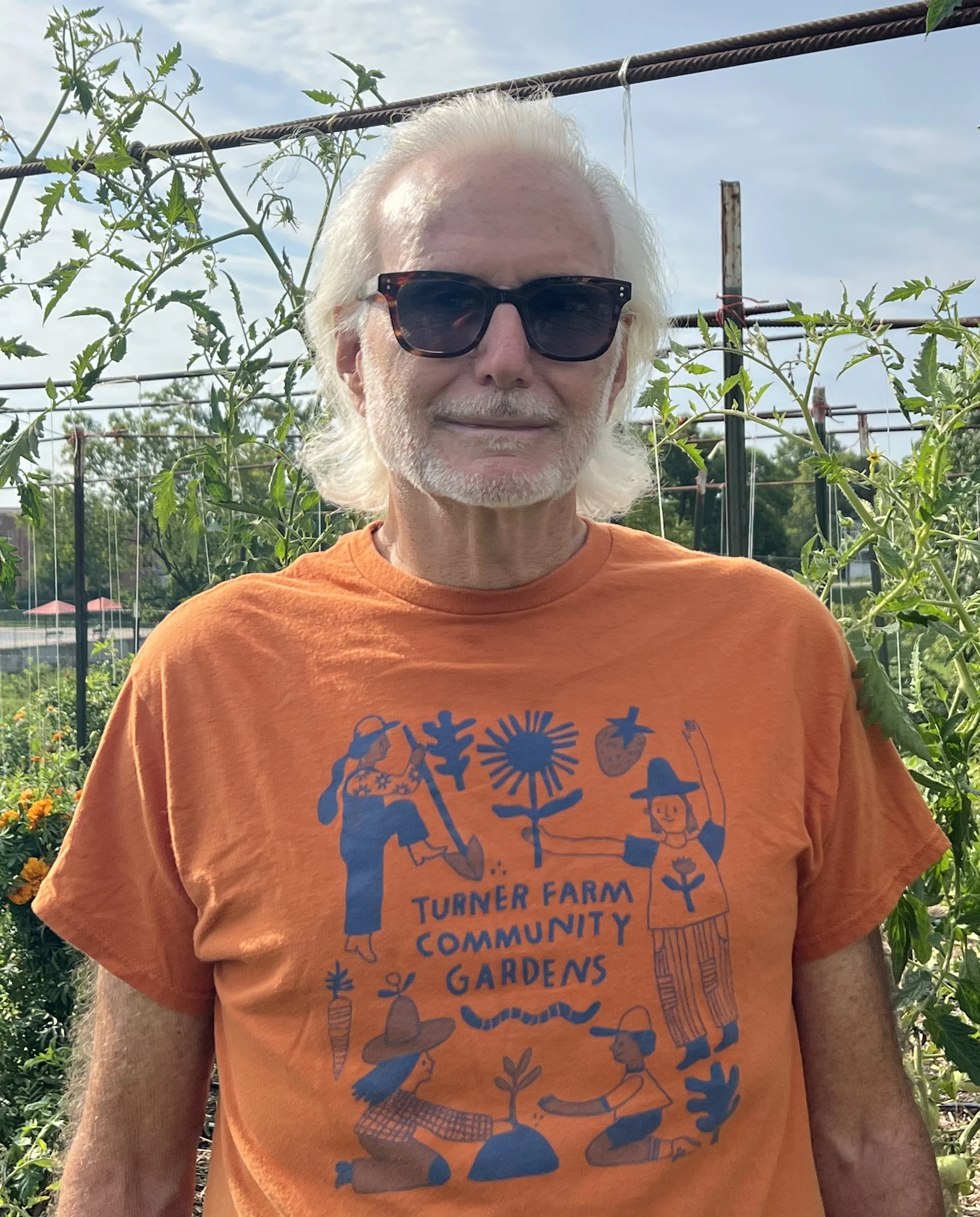 Peter wearing an orange Turner Farm Community Gardens shirt standing in front of plants
