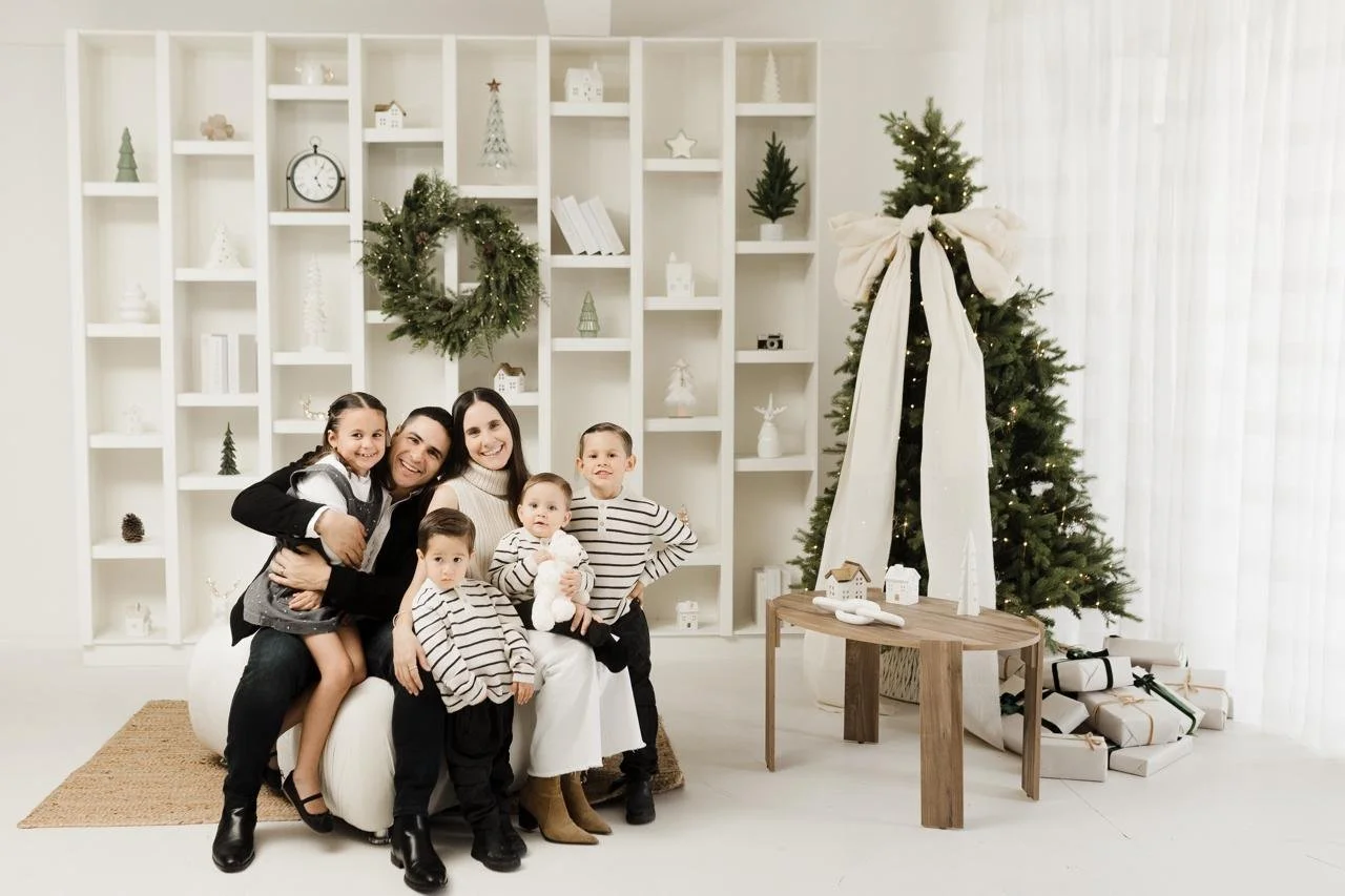 Familia posando en una sala decorada por Navidad, con árbol, regalos y adornos blancos.