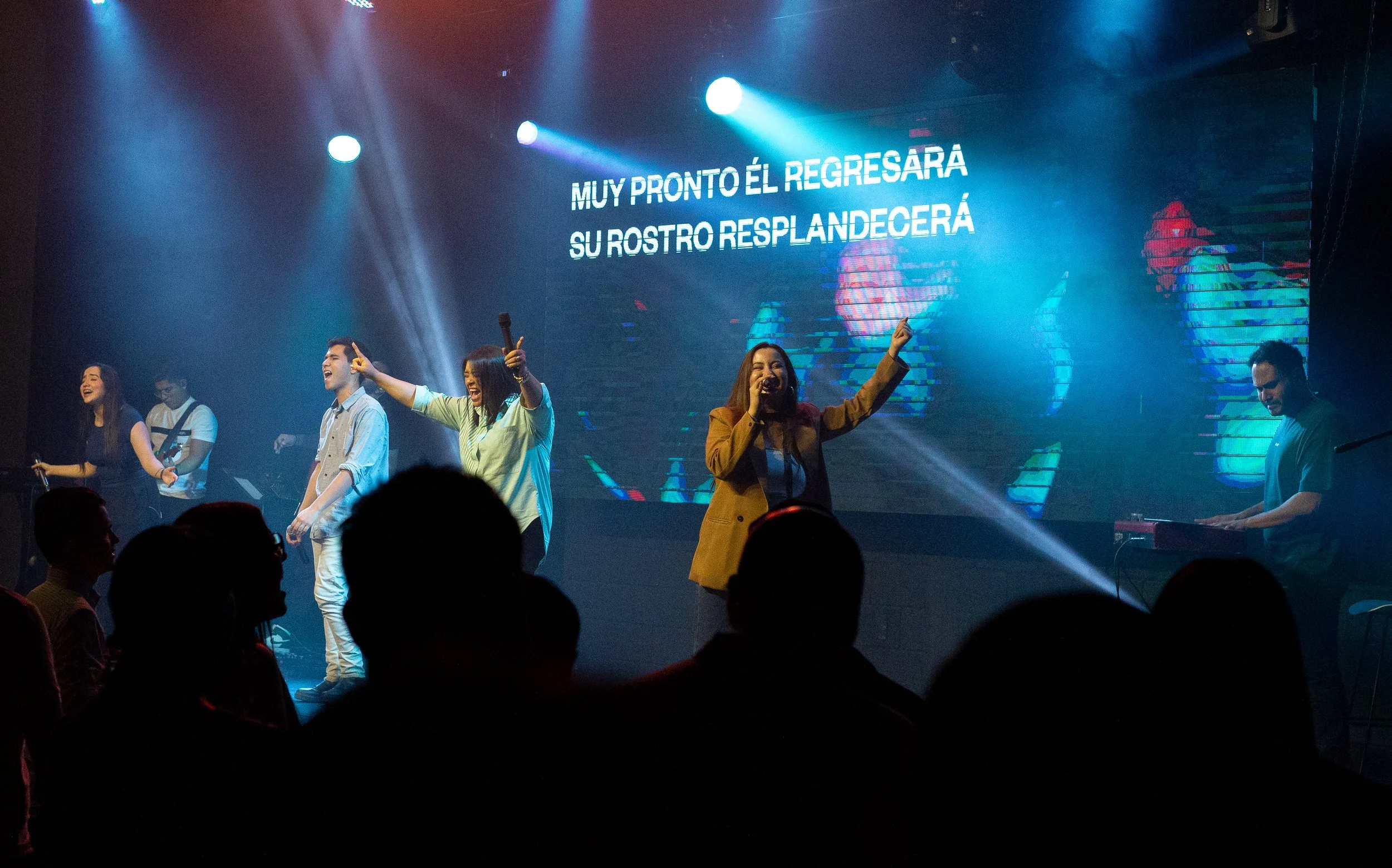 Un grupo de personas cantando y tocando música en un escenario con pantallas de fondo que muestran letras en español, durante una presentación en vivo.
