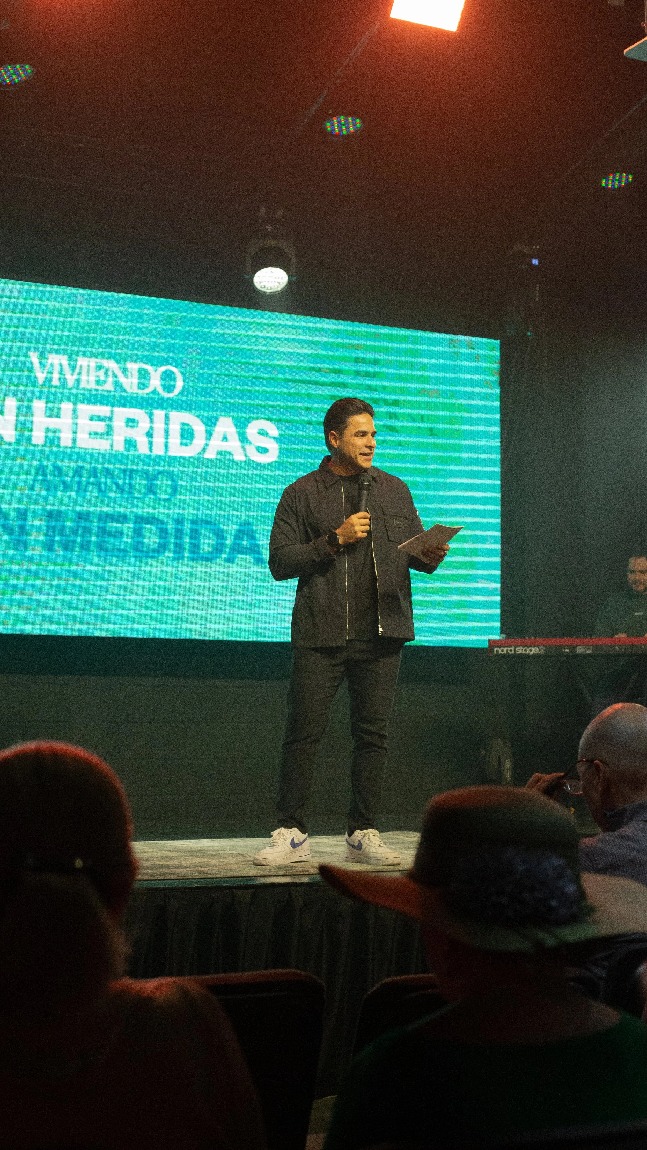 Hombre en el escenario hablando con micrófono, fondo con pantalla digital, audiencia en primer plano, ambiente de concierto o conferencia.