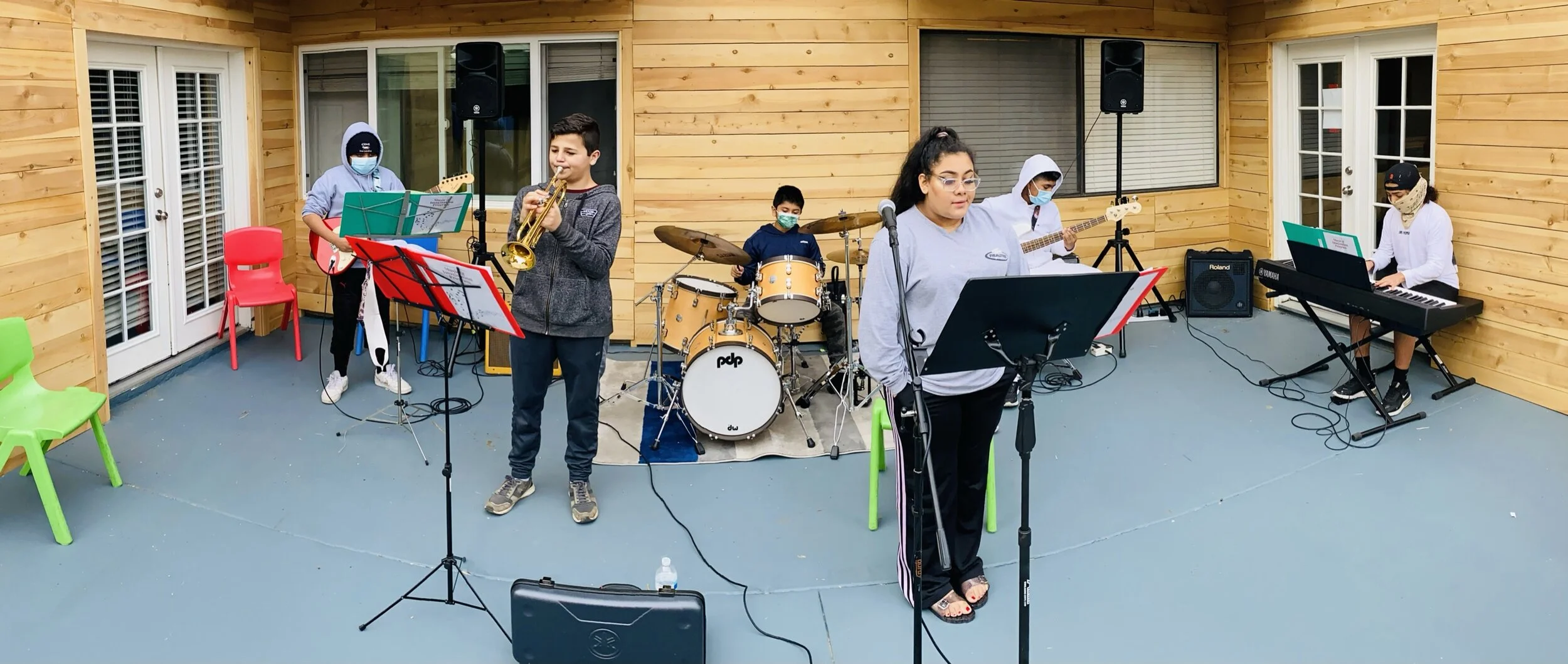 A group of children performing a music concert outdoors on a patio with wooden walls. The band includes a drummer, two keyboard players, a trumpet player, and a bass guitarist. All are wearing face masks, and some are standing while others are seated