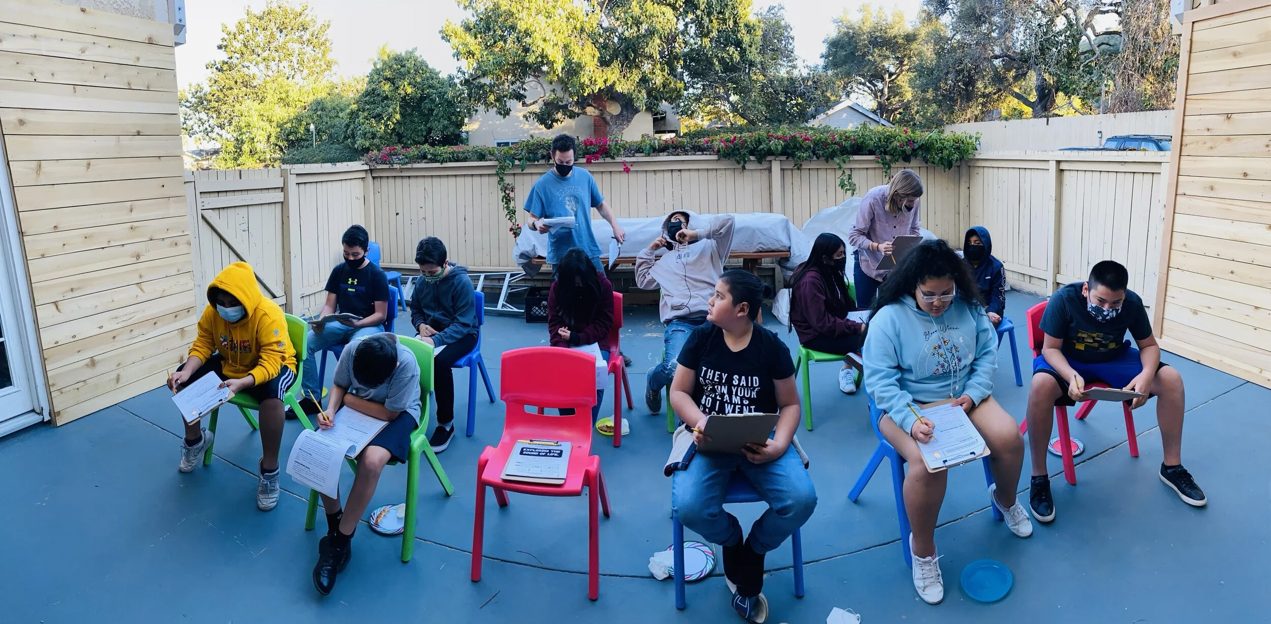 A group of children and a few adults wearing masks seated on colorful chairs outdoors, engaged in an activity with tablets and papers, on a patio enclosed by a wooden fence, with plants and trees in the background.