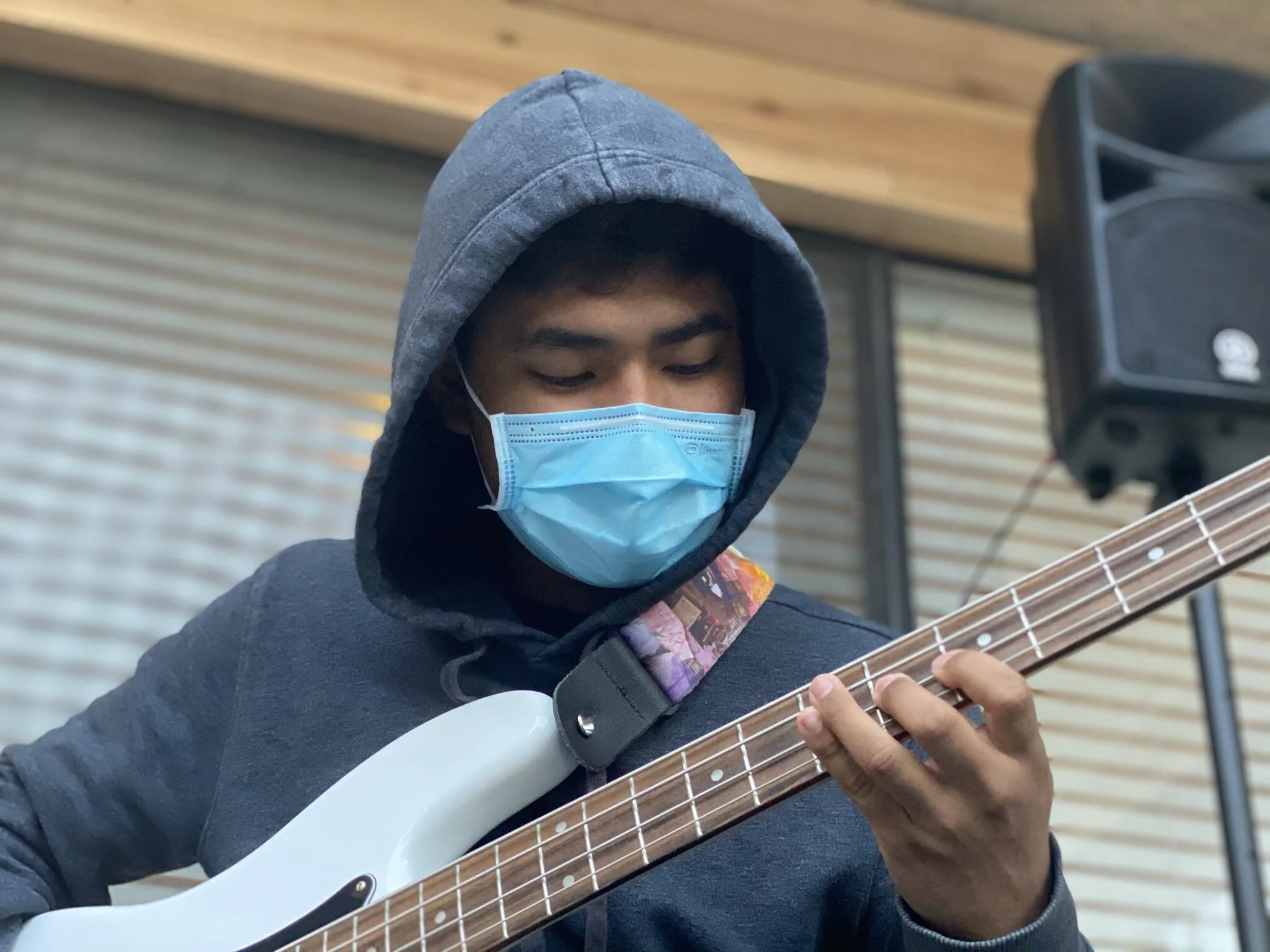 A young man wearing a blue face mask and a gray hoodie, playing an electric bass guitar.