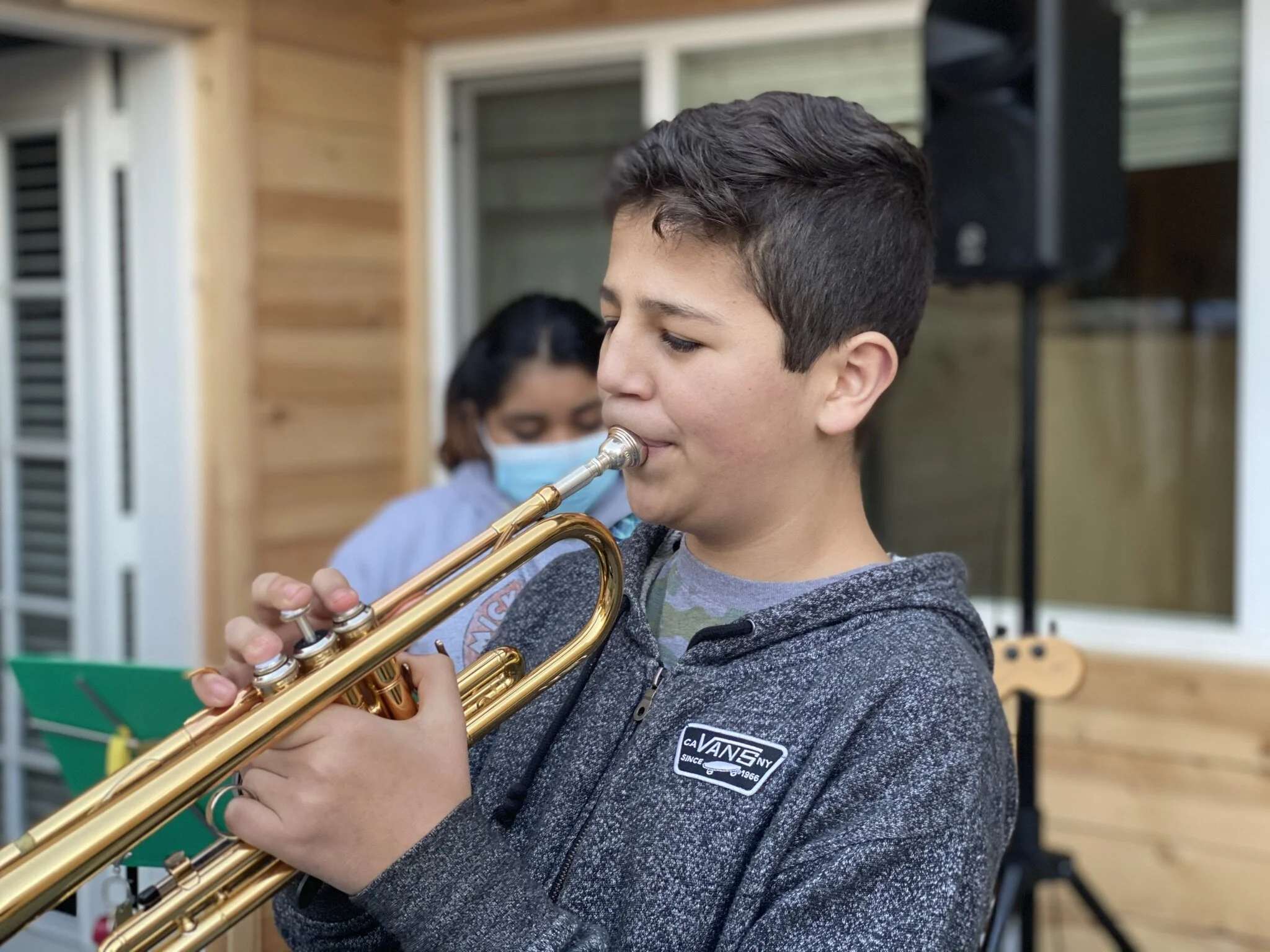 Young boy playing a trumpet with a woman in the background wearing a face mask