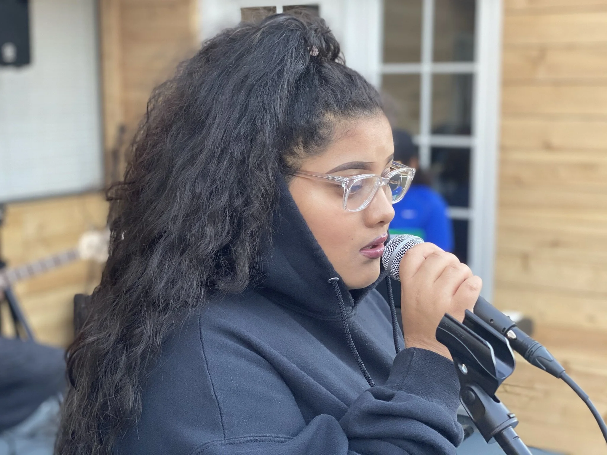 A young woman with long curly dark hair, glasses, and dressed in a black hoodie, speaking into a microphone in an indoor setting with wooden walls.