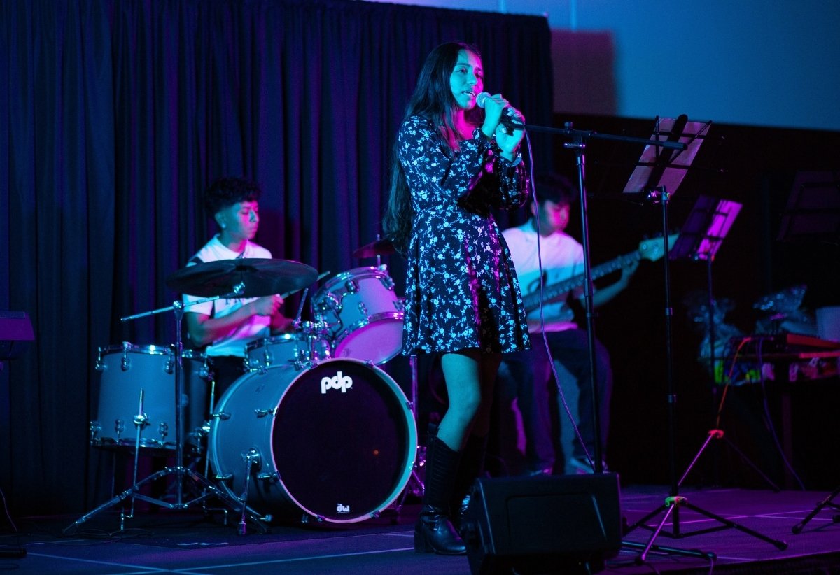 A girl singing into a microphone on stage with two musicians playing instruments behind her, a drummer and a guitarist, in a performance setting with dark curtains and colored stage lighting.