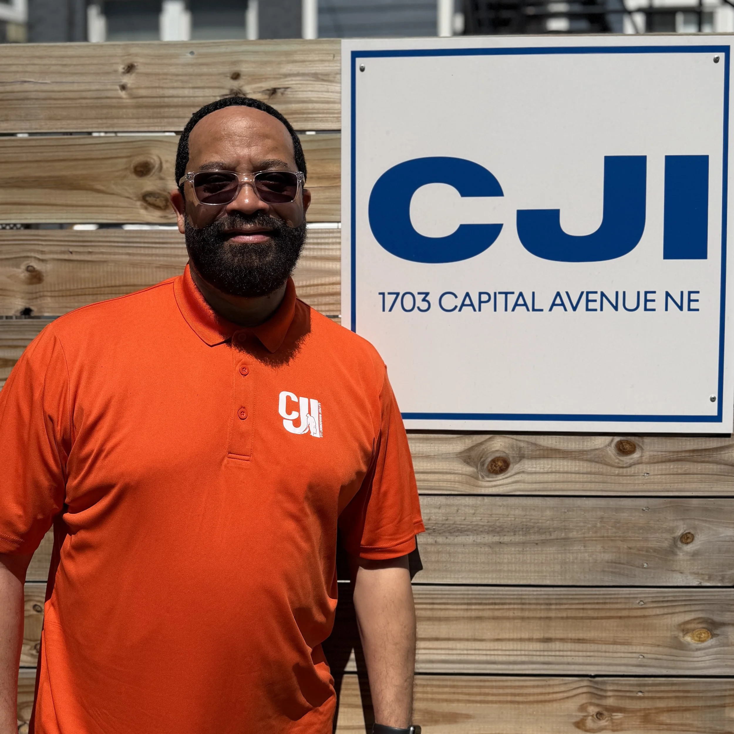 A smiling man wearing sunglasses and a navy blue polo shirt with a logo that reads 'CJI Pest Management' standing in front of a wooden wall and a sign that says 'CJI 1703 Capital Avenue NE'.