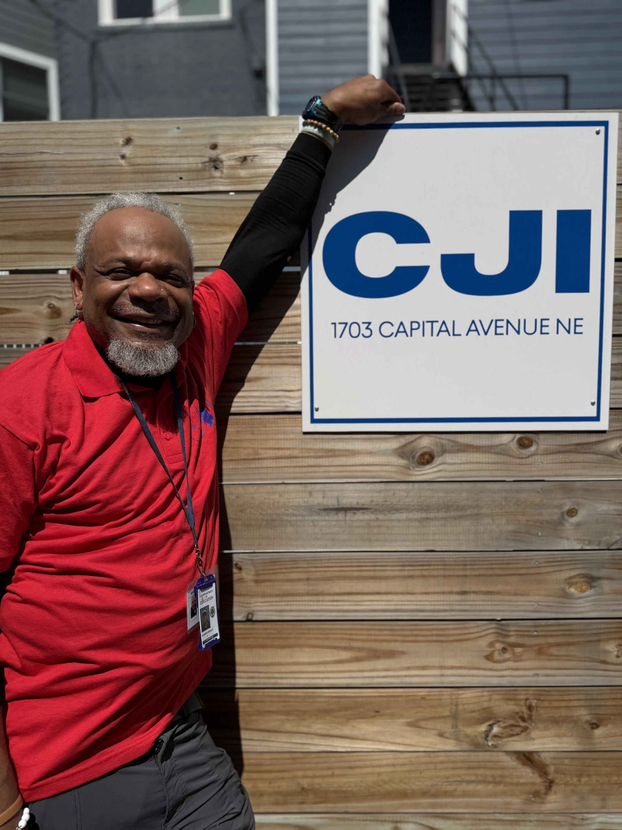 A smiling man wearing a gray polo shirt with with logo and red text, standing outdoors in front of a wooden fence and a sign with blue elements.