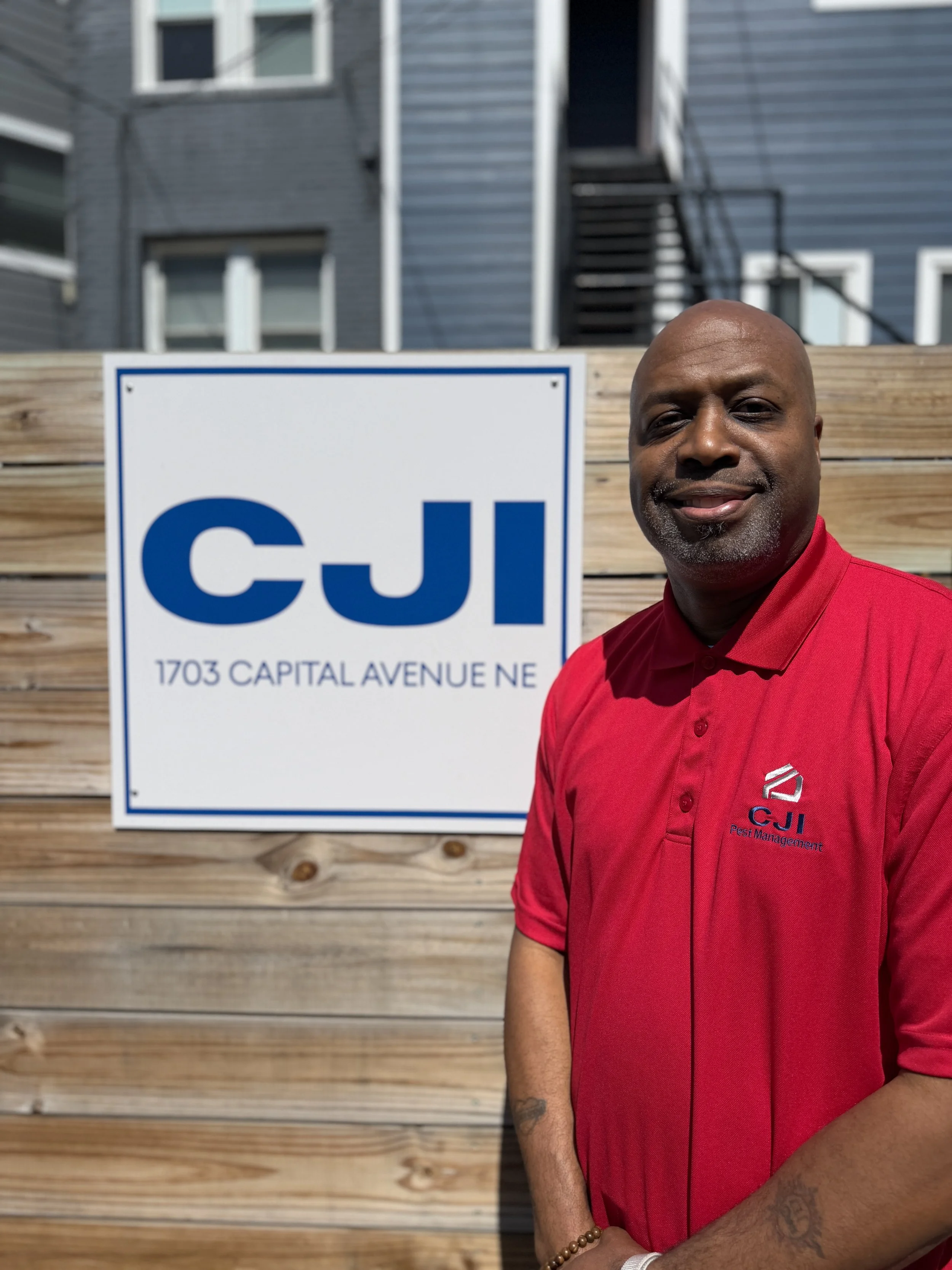 A smiling man with a bald head and a goatee, wearing a gray polo shirt with a logo and text, standing in front of a wooden wall.