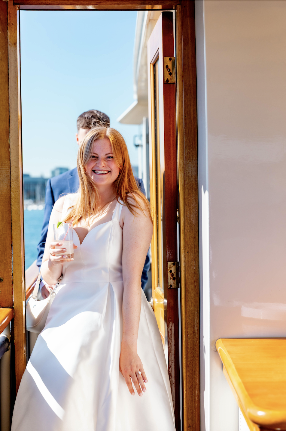 A photo of Hayley, a fiancee, walking towards the camera with a drink in hand, smiling very brightly!