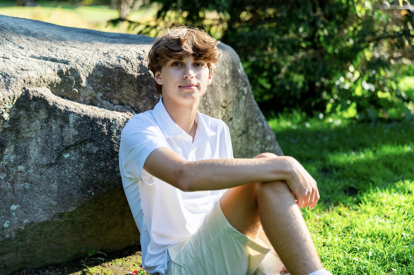 A young person with brown hair and fair skin, wearing a white shirt and shorts, sitting on the grass near a large rock in a park-like setting with green trees and sunlight.